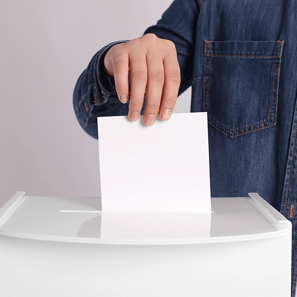 Hand wirft Stimmzettel in eine weiße Wahlurne als Symbol für Wahlbeteiligung